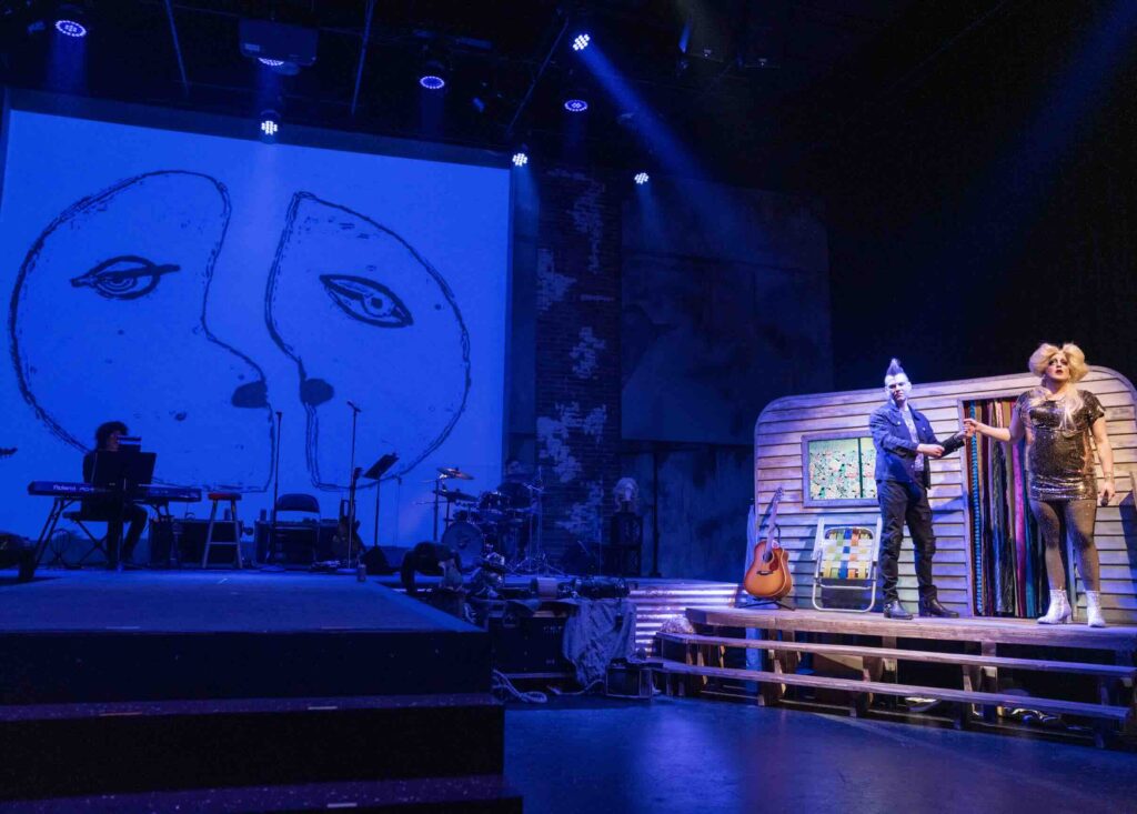A stage set for "Hedwig and the Angry Inch" musical. On the left, a large blue screen displays a sketch of a face with prominent eyes. Musical instruments are set up in front of it. To the right, two actors stand on a small stage made to look like a trailer or mobile home. One actor, likely portraying Hedwig, wears a blonde wig and sparkly dress. The other has a punk-style mohawk. Props include a guitar and a lawn chair. The backdrop shows exposed brick walls, and theatrical lighting bathes the scene in blue. 
